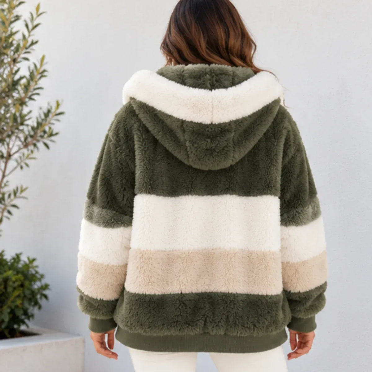 A woman viewed from behind wearing a plush hooded jacket with wide horizontal stripes in olive green, cream, and beige, standing against a light gray wall with a plant at the side.