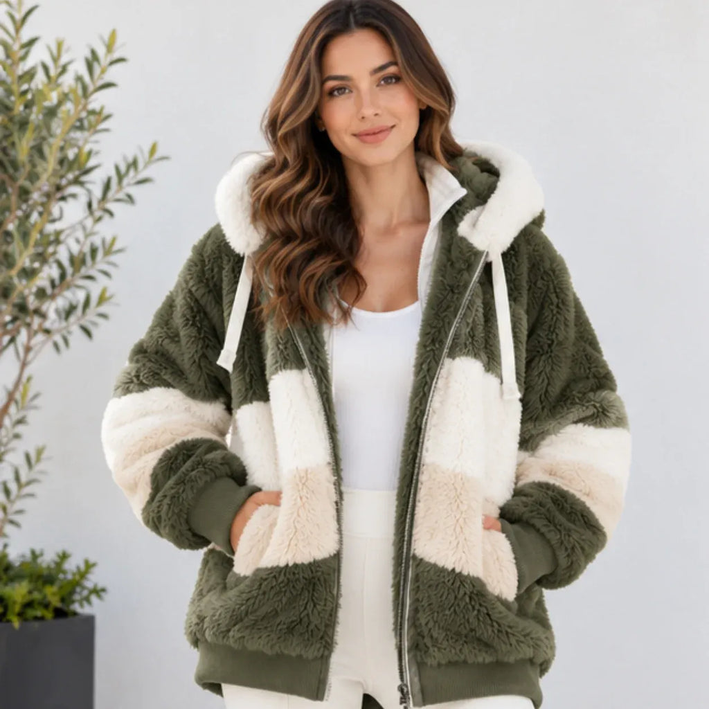 A woman wearing a plush zip-up hoodie jacket with a color-blocked design in olive green and cream, styled with a white tank top and white pants against a light gray background.