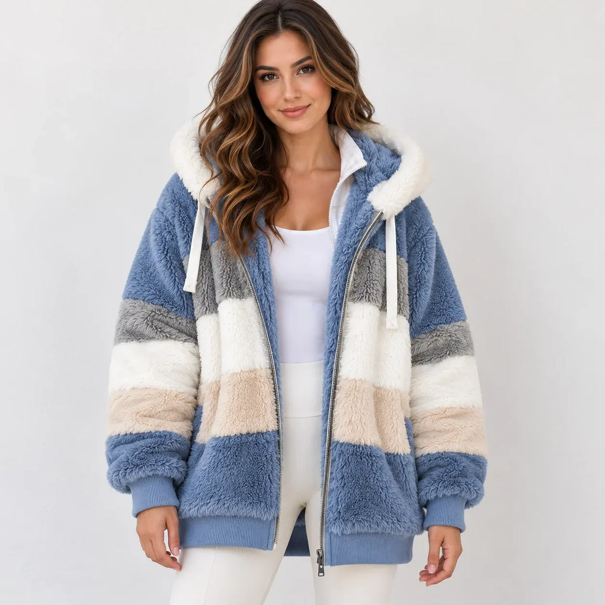 A woman wearing a plush hooded zip-up jacket with horizontal stripes in dusty blue, cream, beige, and gray, paired with a white tank top and white pants, against a light gray studio background.
