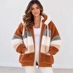 A woman wearing a plush zip-up hooded jacket with horizontal color-blocked stripes in rust brown, cream, and gray, styled over a white top and white pants against a clean white studio background.