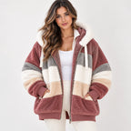 A woman wearing a plush sherpa zip-up hoodie jacket with horizontal stripes in dusty rose, cream, beige, and gray, styled over a white tank top and white pants against a light gray studio background.