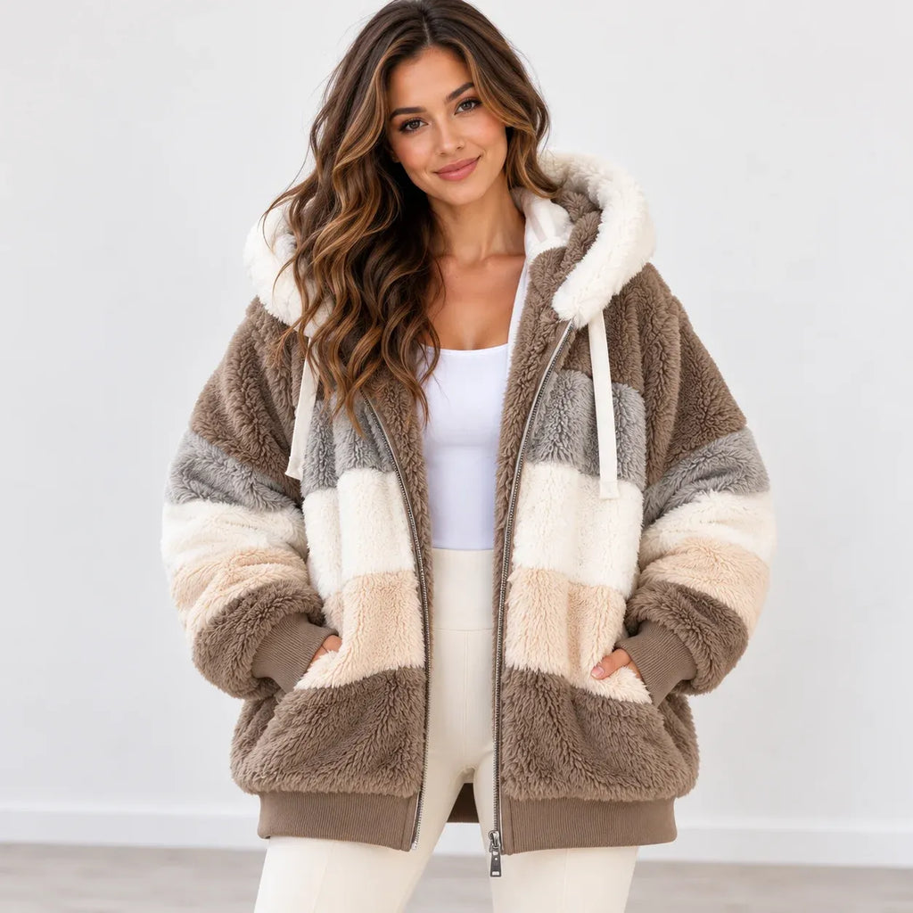 A woman wearing a plush teddy-style zip-up jacket with large color-blocked panels in taupe brown, cream, and light gray, paired with a white tank top and white pants against a clean light gray studio background.
