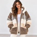 A woman wearing a plush teddy-style zip-up jacket with large color-blocked panels in taupe brown, cream, and light gray, paired with a white tank top and white pants against a clean light gray studio background.
