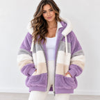 A woman wearing a chunky, oversized sherpa fleece zip-up hoodie jacket with horizontal color-blocked stripes in lavender purple, cream, and light gray. The jacket features a white sherpa-lined hood with drawstrings, a front zipper, and side pockets, styled over a white tank top and white pants.