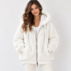 A woman wearing a plush white sherpa zip-up hoodie jacket with a drawstring hood and front pockets, photographed against a clean light gray studio background.