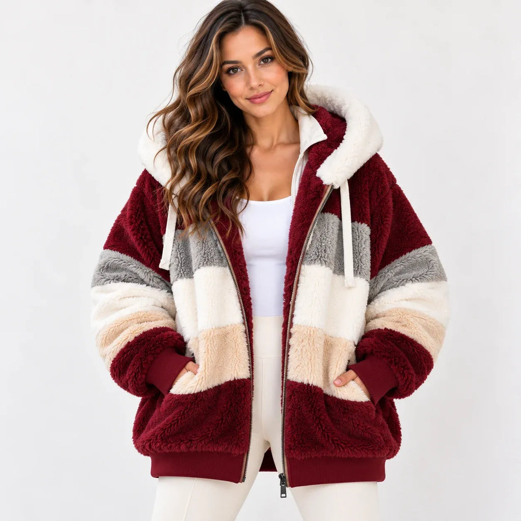 A woman wearing a plush zip-up hooded jacket with block stripes in burgundy, cream, and gray, paired with a white tank top and white pants, against a plain light gray background.
