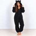 A woman stands barefoot wearing a black plush bear onesie with a hood and ear details, posed against a plain white studio background.