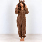 A woman stands barefoot wearing a brown plush hooded one-piece pajama with bear ears on the hood, photographed against a plain light gray wall and pale wood floor.