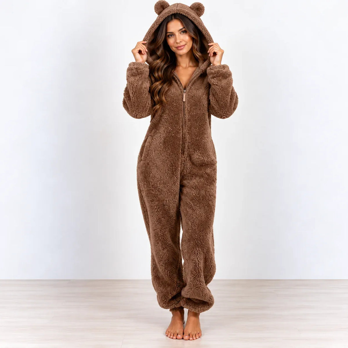 A woman stands barefoot wearing a brown plush hooded one-piece pajama with bear ears on the hood, photographed against a plain light gray wall and pale wood floor.