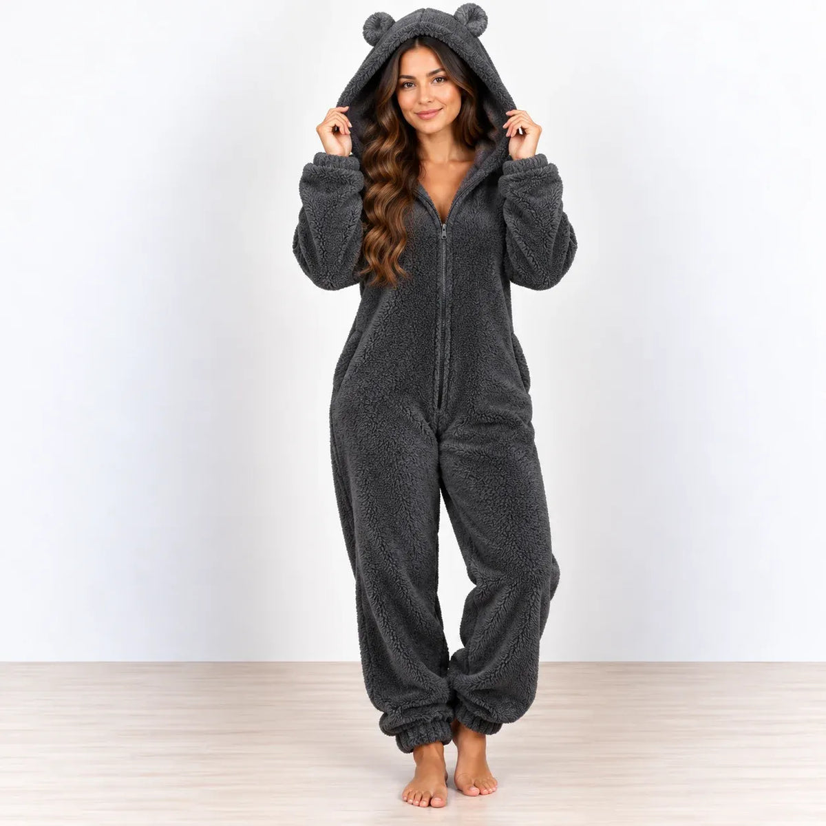 A woman stands barefoot wearing a dark gray plush zip-up onesie with a bear-ear hood, posed against a plain white wall and light wood floor.