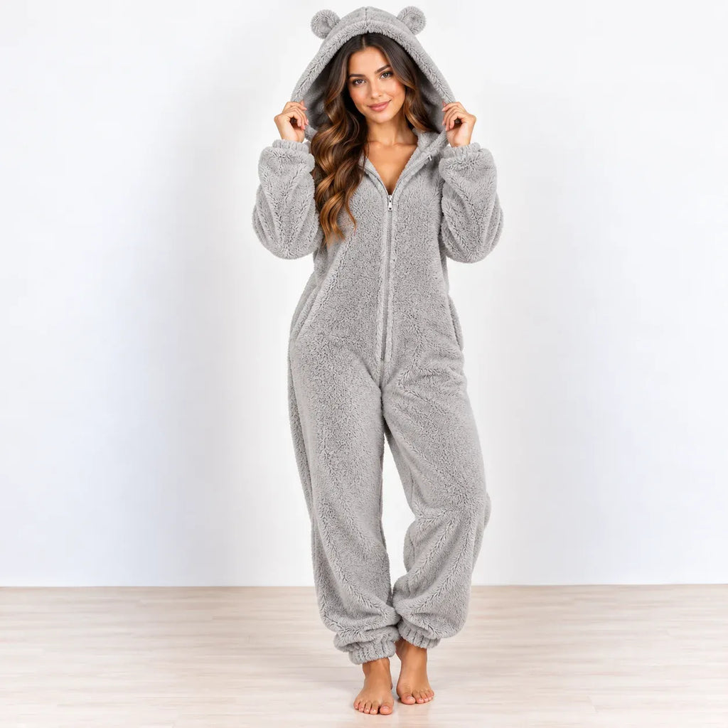 A woman wearing a light gray plush hooded one-piece pajama with bear ears on the hood, standing barefoot against a plain white wall and light wood floor.