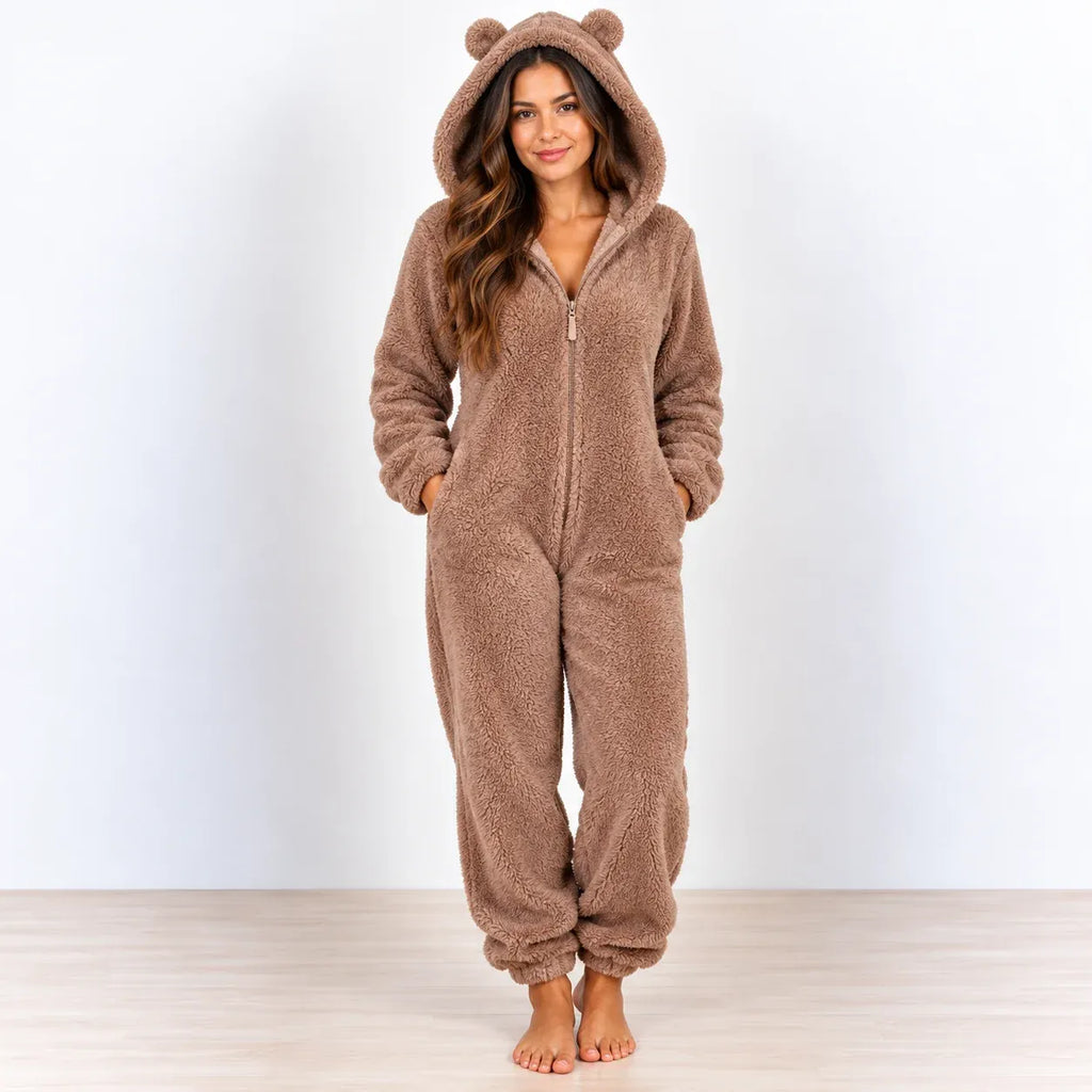 A woman stands barefoot wearing a plush brown zip-up hooded onesie with bear ears on the hood, photographed against a plain light wall and pale wood floor.