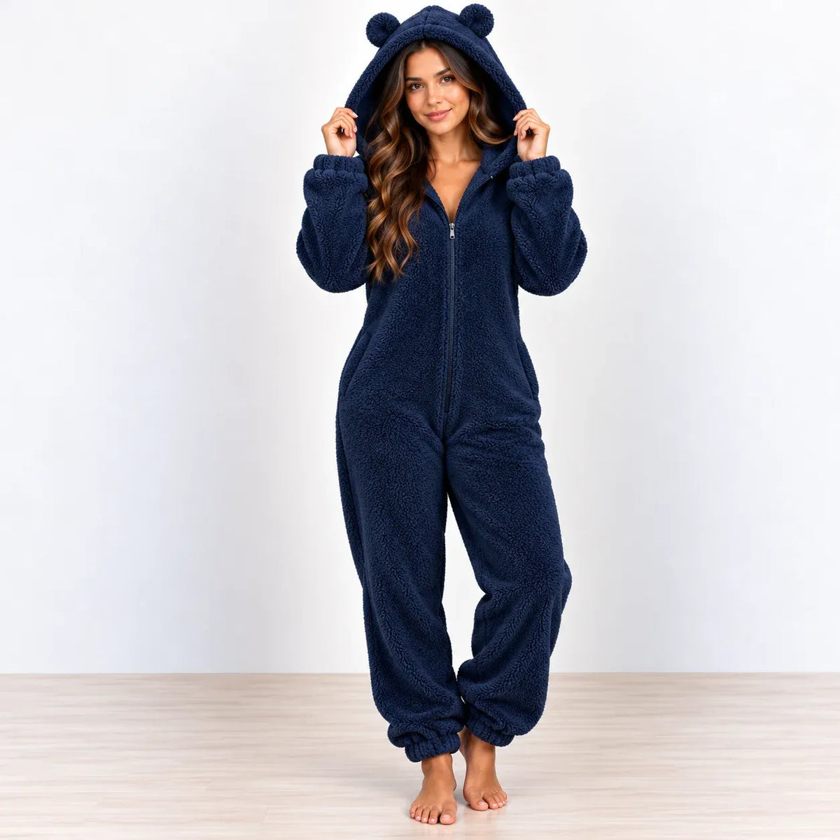 A woman stands barefoot in a studio wearing a navy blue plush one-piece pajama with a full zip front, long sleeves, and a hood with two bear ears.