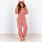 A woman wearing a plush, one-piece teddy bear onesie in dusty rose pink, standing against a white wall and light wood floor.