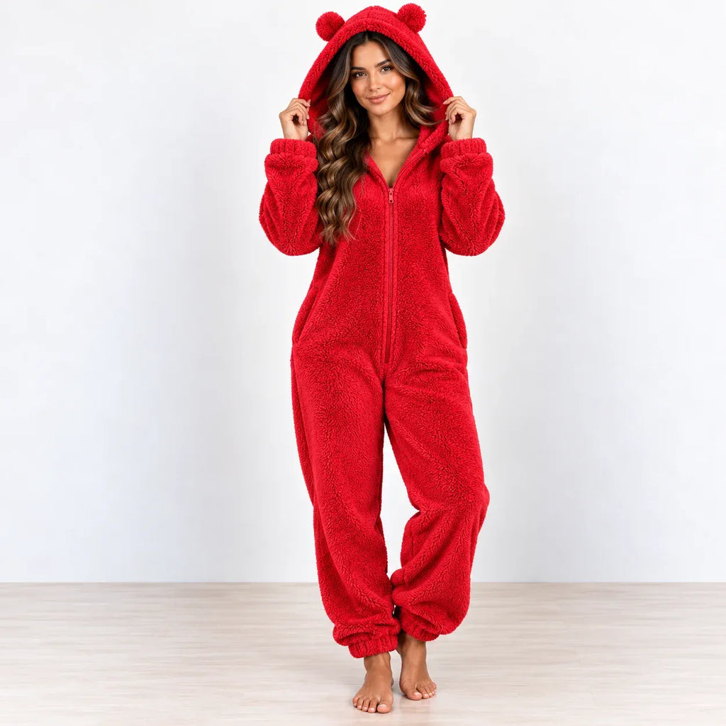A woman wearing a plush red one-piece pajama suit with a hood and bear-ear pom-poms, standing barefoot against a plain white wall and light wood floor.