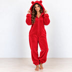 A woman wearing a plush red one-piece pajama suit with a hood and bear-ear pom-poms, standing barefoot against a plain white wall and light wood floor.