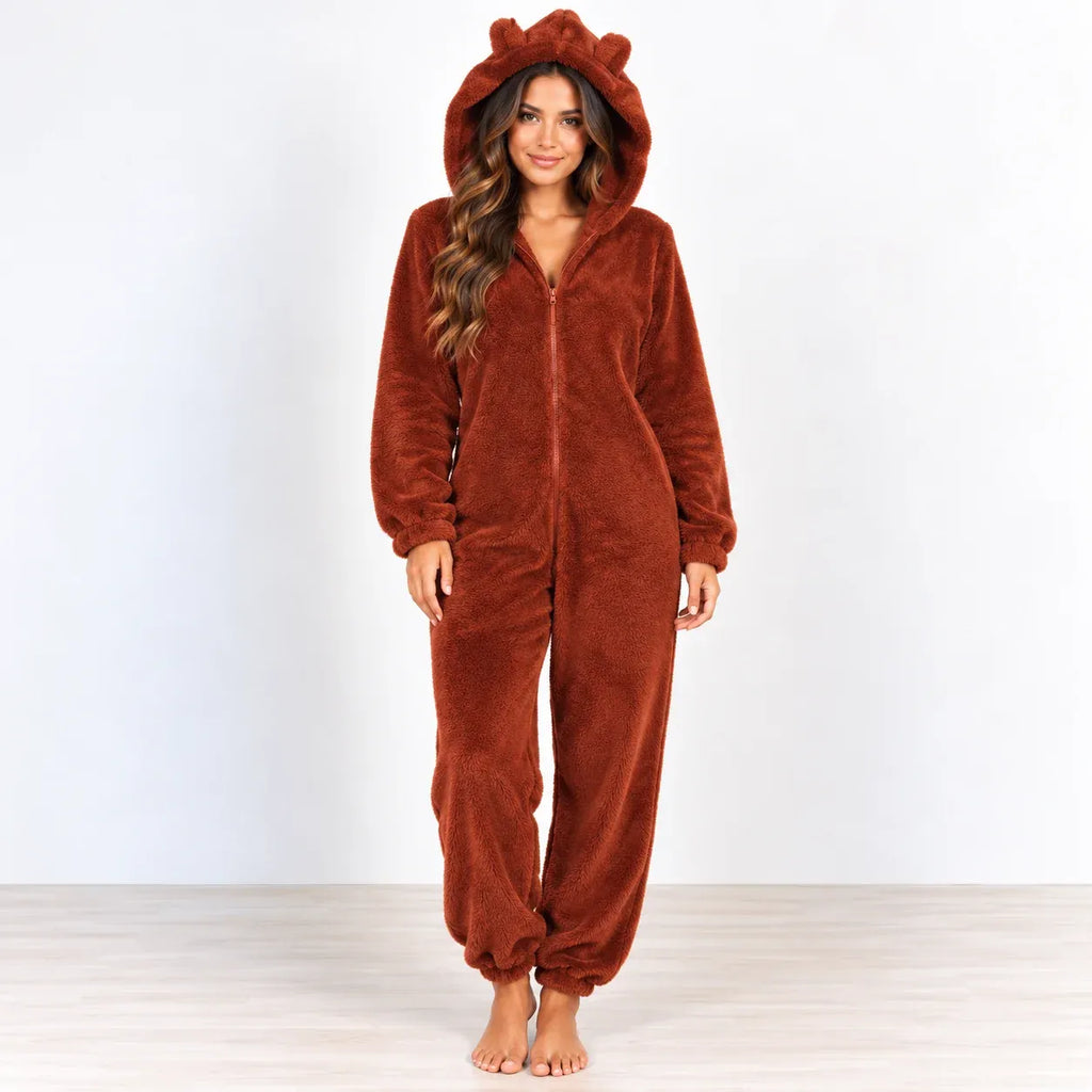 A woman wearing a plush brown hooded one-piece pajama with bear ears on the hood, standing barefoot against a plain white studio background.