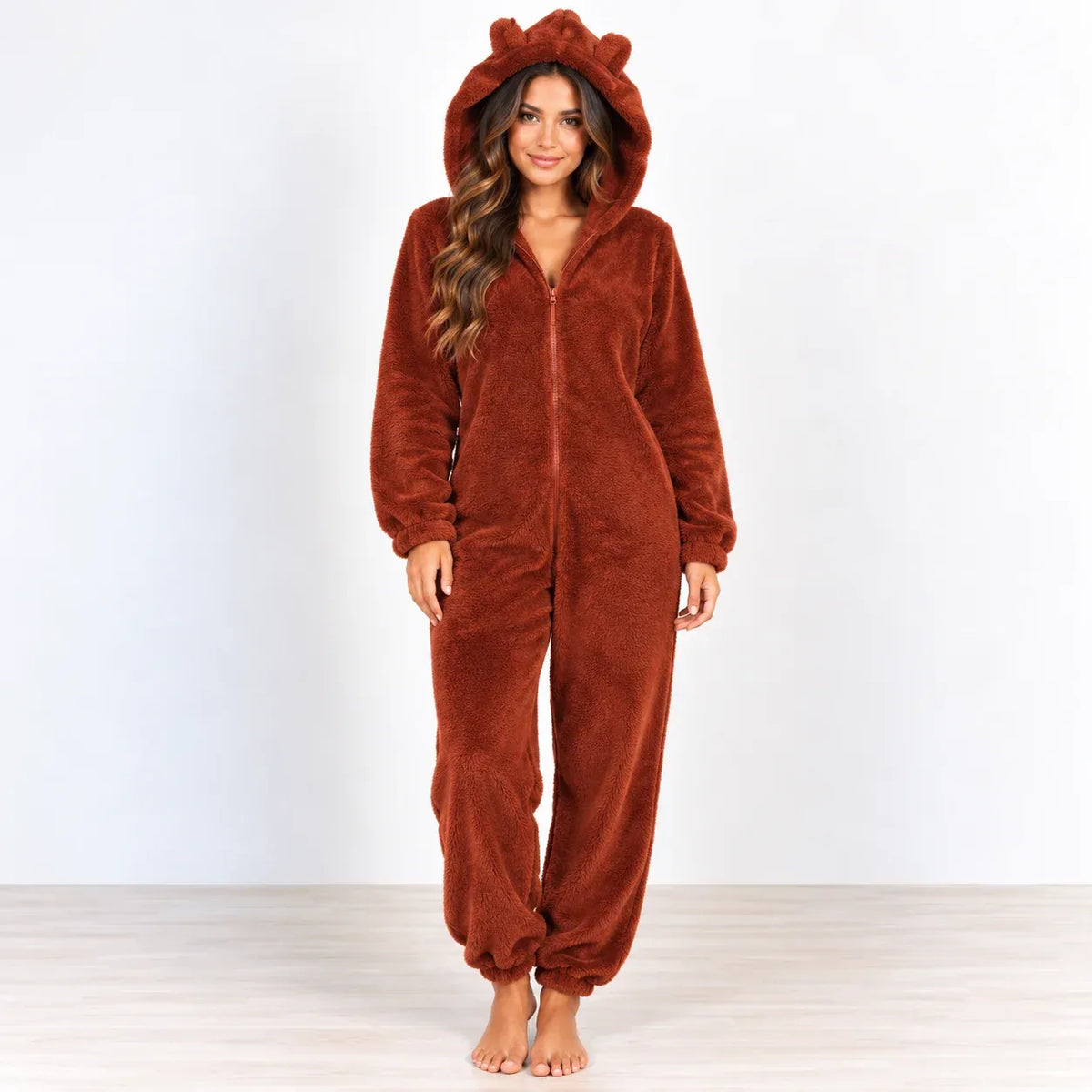 A woman wearing a plush brown hooded one-piece pajama with bear ears on the hood, standing barefoot against a plain white studio background.