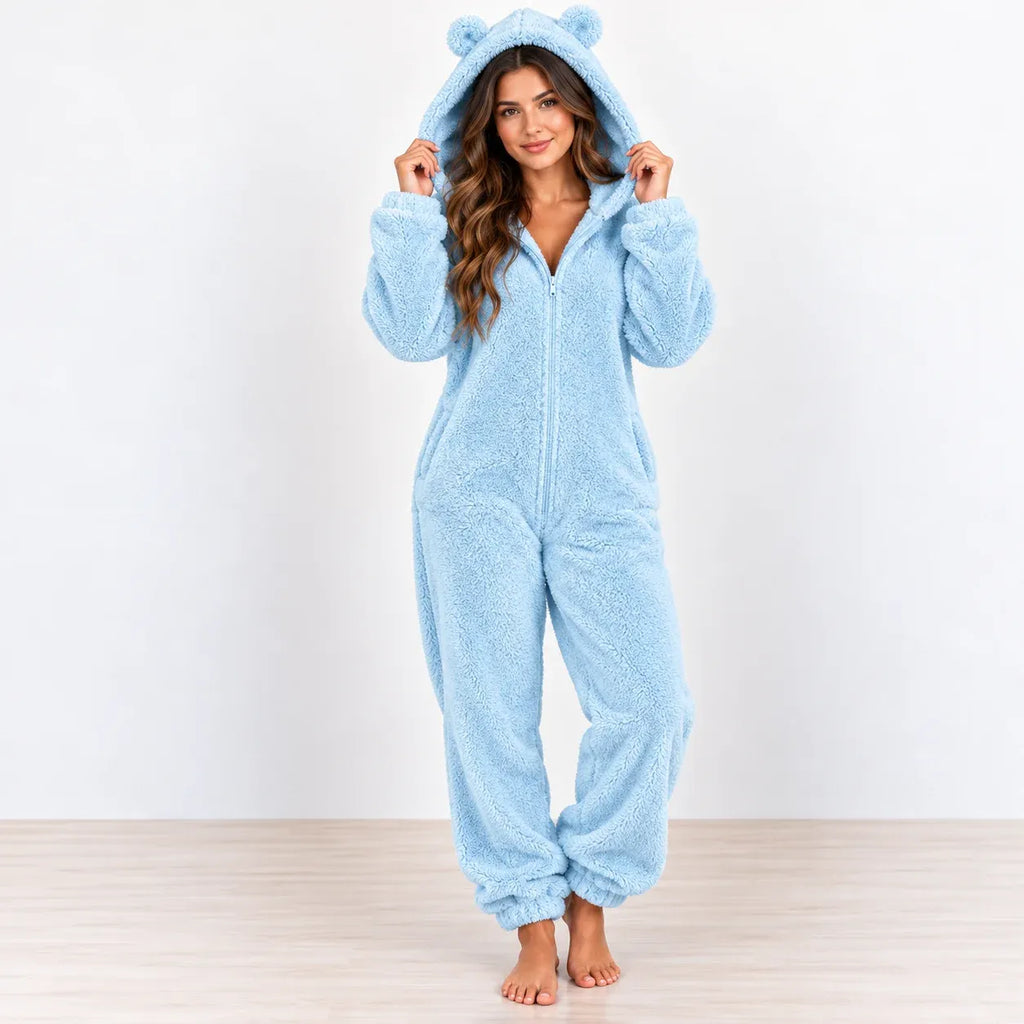 A woman wearing a light blue plush hooded one-piece pajama with bear ears on the hood, standing barefoot against a plain white wall and light wood floor.