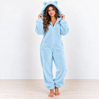 A woman wearing a light blue plush hooded one-piece pajama with bear ears on the hood, standing barefoot against a plain white wall and light wood floor.