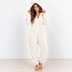 A woman wearing a fluffy cream-colored hooded onesie with bear ears on the hood, standing barefoot against a light gray wall and pale wood floor.
