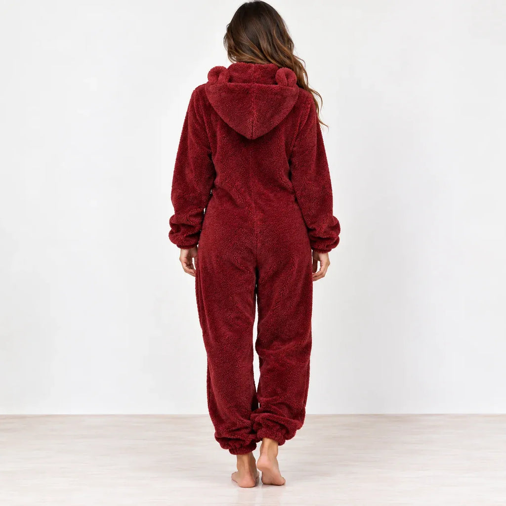 A woman viewed from behind wearing a plush, long-sleeve one-piece pajama in deep burgundy red. The pajama features a hood with faux-fur ear details on the top and cuffed ankles.