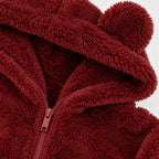 Close-up of a plush teddy fleece zip-up hoodie in deep burgundy red, featuring a hood with two pom-poms and a matching zipper pull.