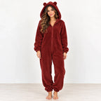 A woman wearing a plush burgundy bear onesie pajama with a hood, ear details, and front zipper, standing against a plain white studio background.