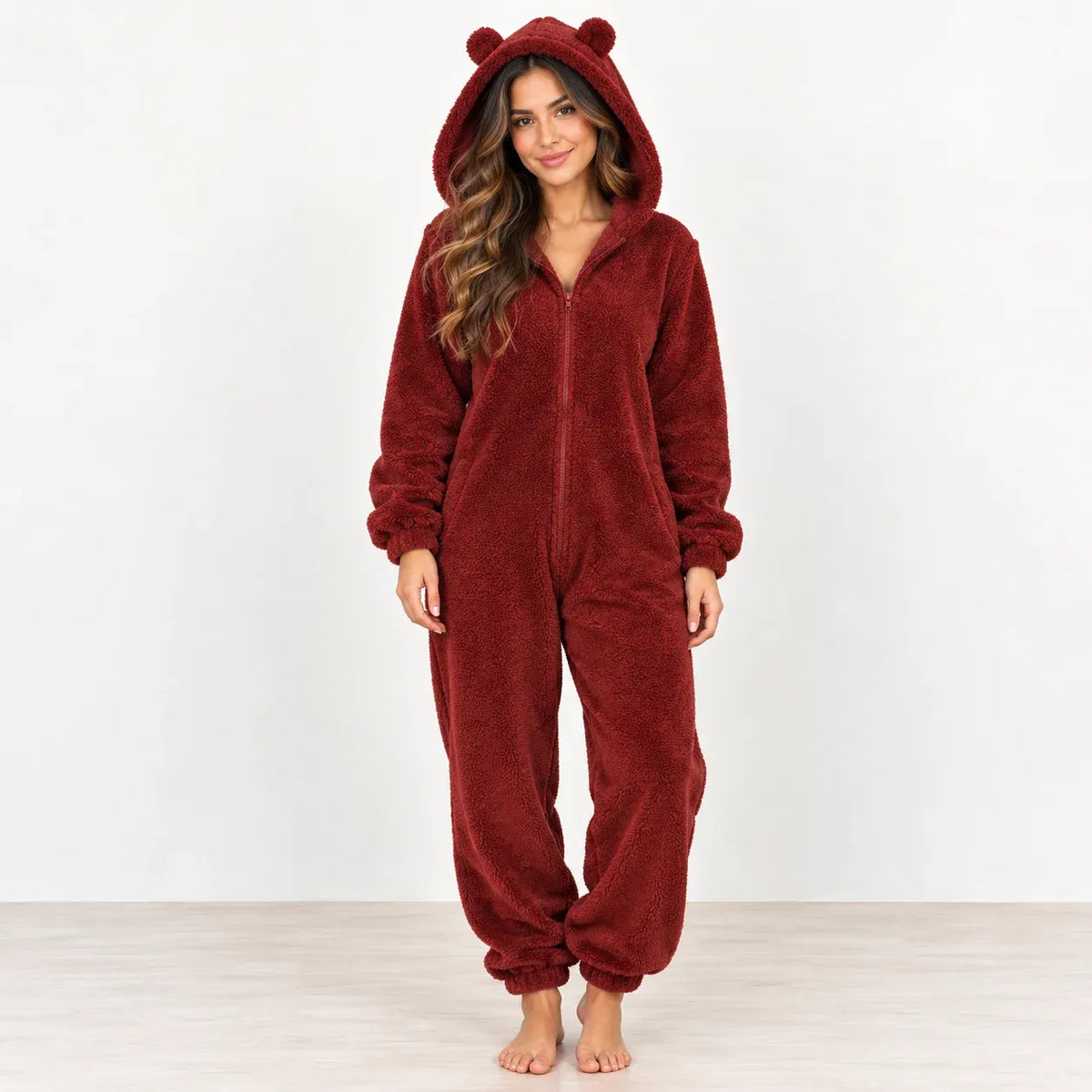 A woman wearing a plush burgundy bear onesie pajama with a hood, ear details, and front zipper, standing against a plain white studio background.