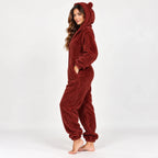 A woman modeling a plush, one-piece bear-themed pajama suit in a deep burgundy red color, standing against a plain white studio background.