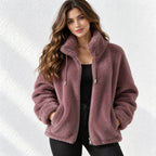 A woman wearing a dusty mauve sherpa fleece zip-up jacket with a high collar, styled over a black top and black pants, standing against a white textured wall.