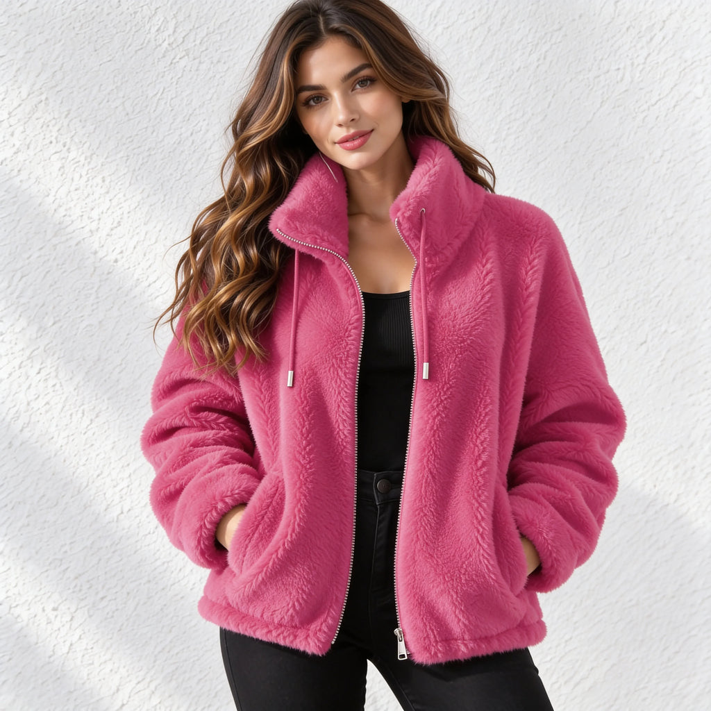 A woman wearing a plush pink sherpa zip-up jacket with a high collar, standing against a textured white wall.