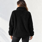 A woman viewed from behind wearing a black sherpa fleece jacket with a high collar, paired with black pants, against a white textured wall.