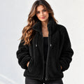 A woman wearing a black sherpa fleece zip-up jacket with a high collar, front zipper, and drawstrings, posed against a light gray textured wall.