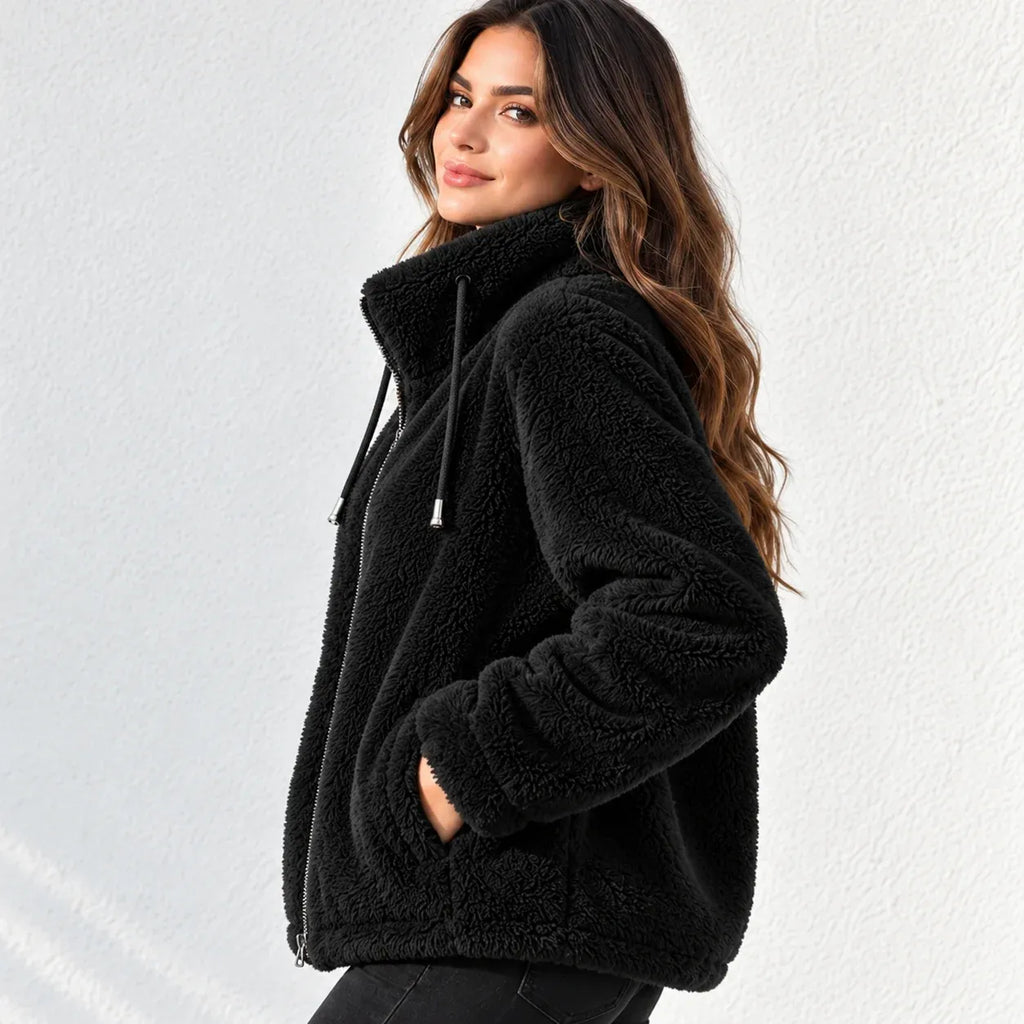 A woman wearing a black sherpa fleece zip-up hoodie jacket with a high collar and drawstrings, posed against a light gray textured wall.