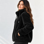 A woman wearing a black sherpa fleece zip-up hoodie jacket with a high collar and drawstrings, posed against a light gray textured wall.
