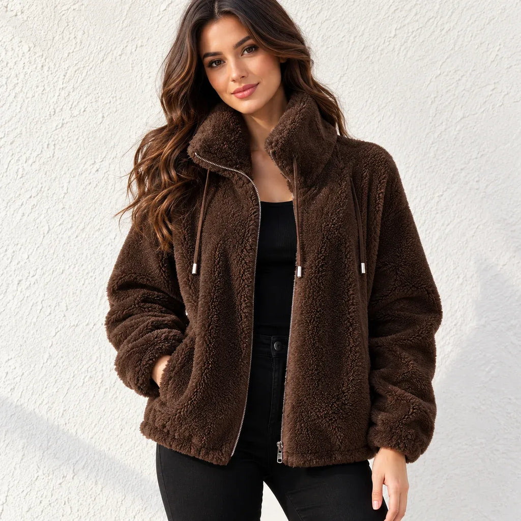 A woman wearing a brown sherpa fleece zip-up jacket with a high collar, standing against a textured white wall.