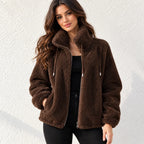 A woman wearing a brown sherpa fleece zip-up jacket with a high collar, standing against a textured white wall.
