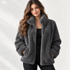 A woman wearing a dark gray sherpa fleece zip-up jacket with a high collar, standing against a textured white wall.