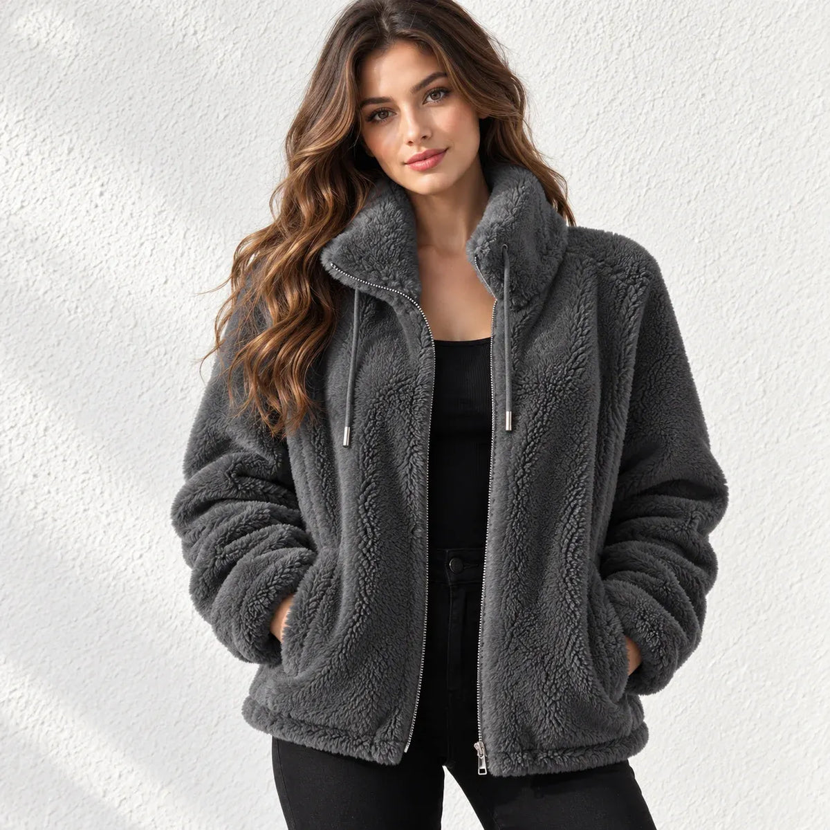 A woman wearing a dark gray sherpa fleece zip-up jacket with a high collar, standing against a textured white wall.