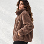 A woman wearing a brown plush fleece jacket with a high collar and drawstrings, standing against a white textured wall.