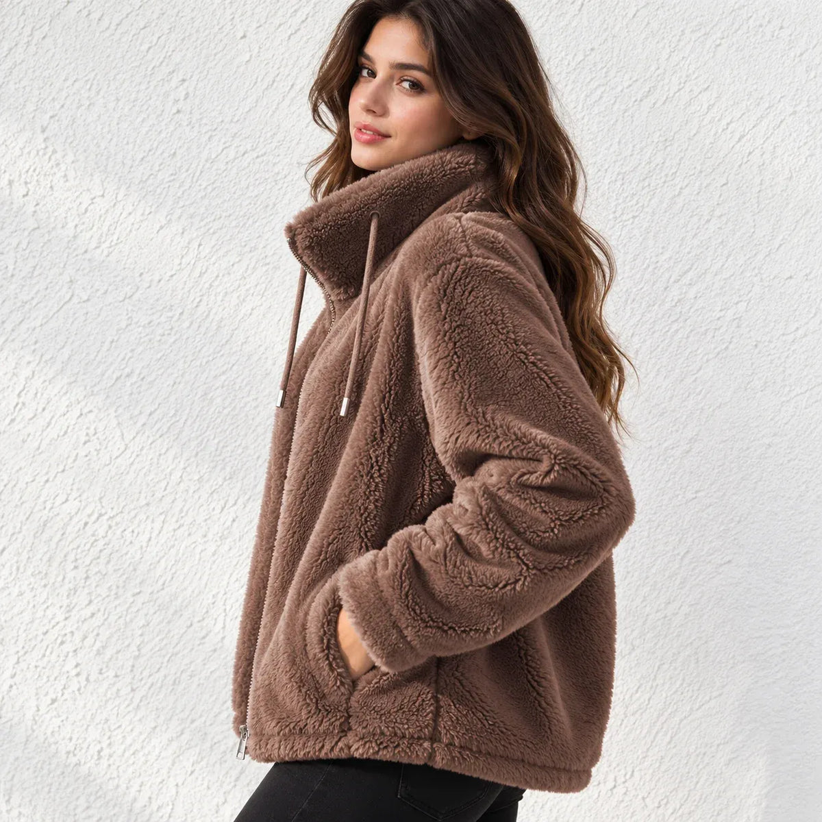 A woman wearing a brown plush fleece jacket with a high collar and drawstrings, standing against a white textured wall.