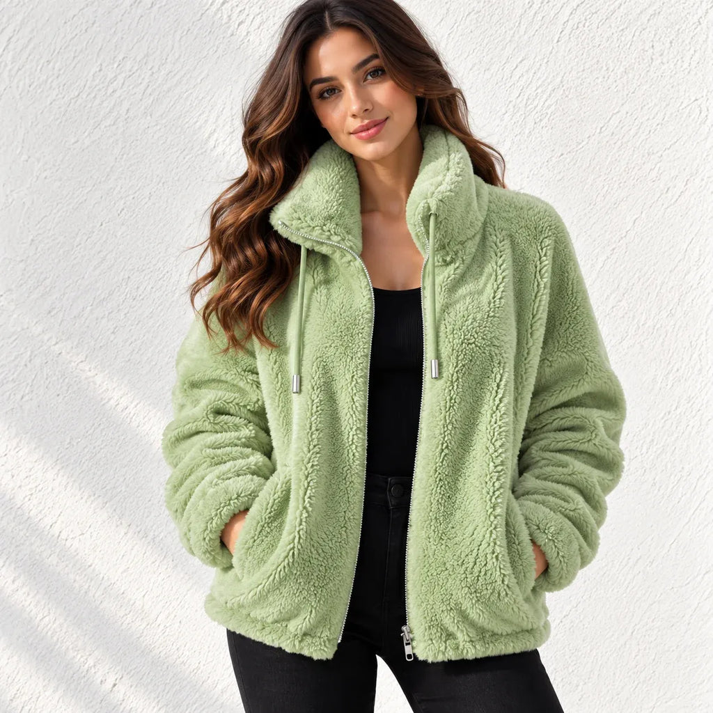A woman wearing a light sage green sherpa fleece zip-up jacket with a high collar, styled over a black top and black pants, standing against a white textured wall.
