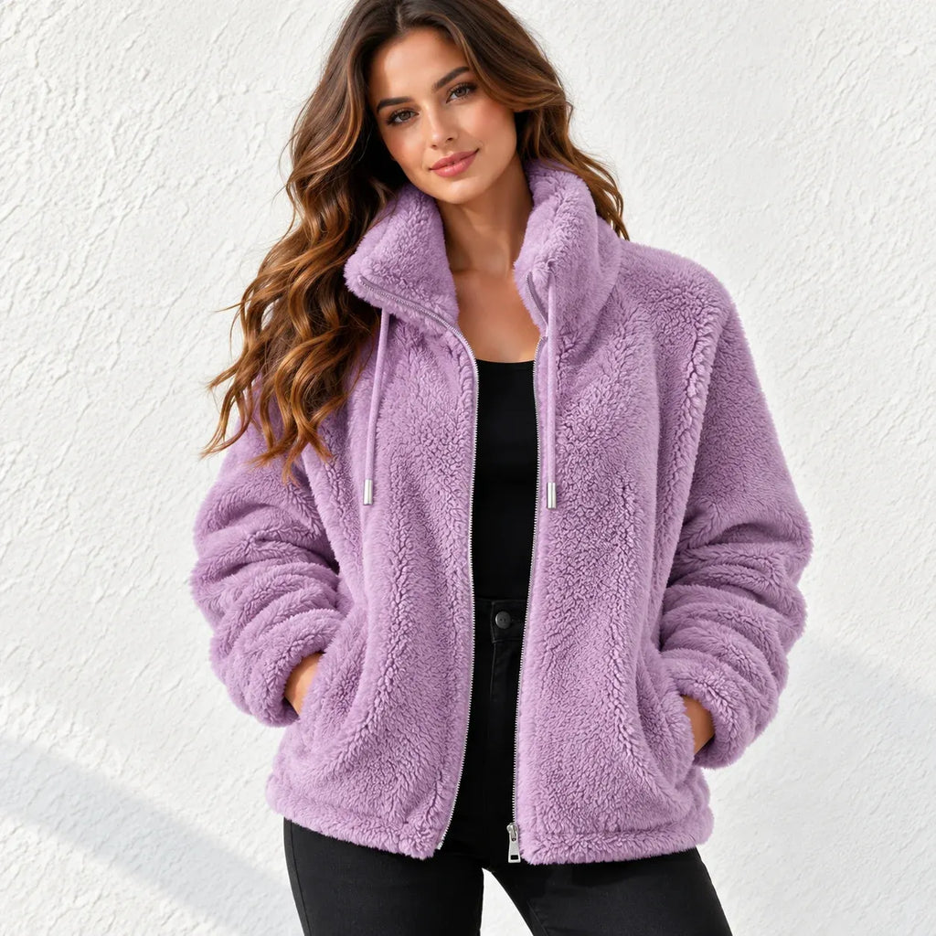 A woman wearing a lavender purple sherpa fleece zip-up jacket with a high collar and drawstrings, paired with a black top and black pants, standing against a white textured wall.