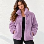A woman wearing a lavender purple sherpa fleece zip-up jacket with a high collar and drawstrings, paired with a black top and black pants, standing against a white textured wall.