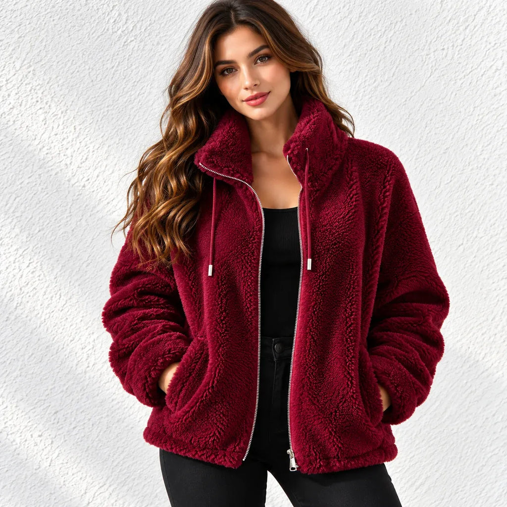 A woman wearing a plush, deep burgundy sherpa fleece zip-up jacket with a high collar, standing against a textured white wall.