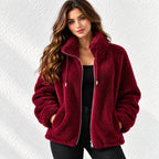 A woman wearing a plush, deep burgundy sherpa fleece zip-up jacket with a high collar, standing against a textured white wall.