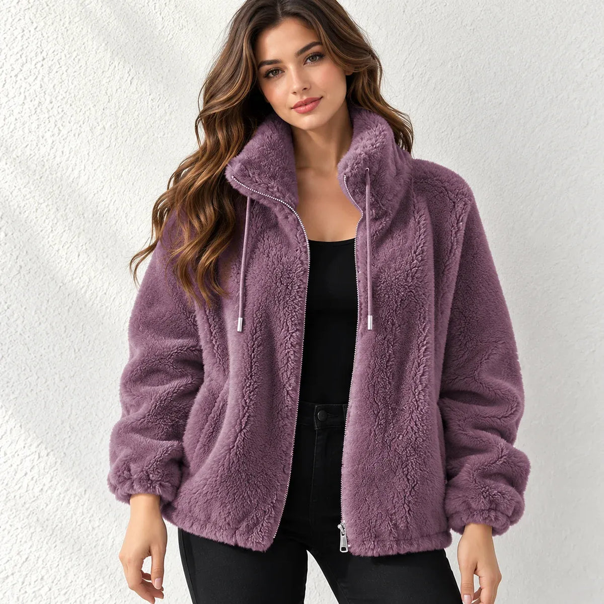 A woman wearing a plush, high-collar fleece jacket in a dusty mauve-purple color, styled over a black top and black pants, standing against a textured white wall.