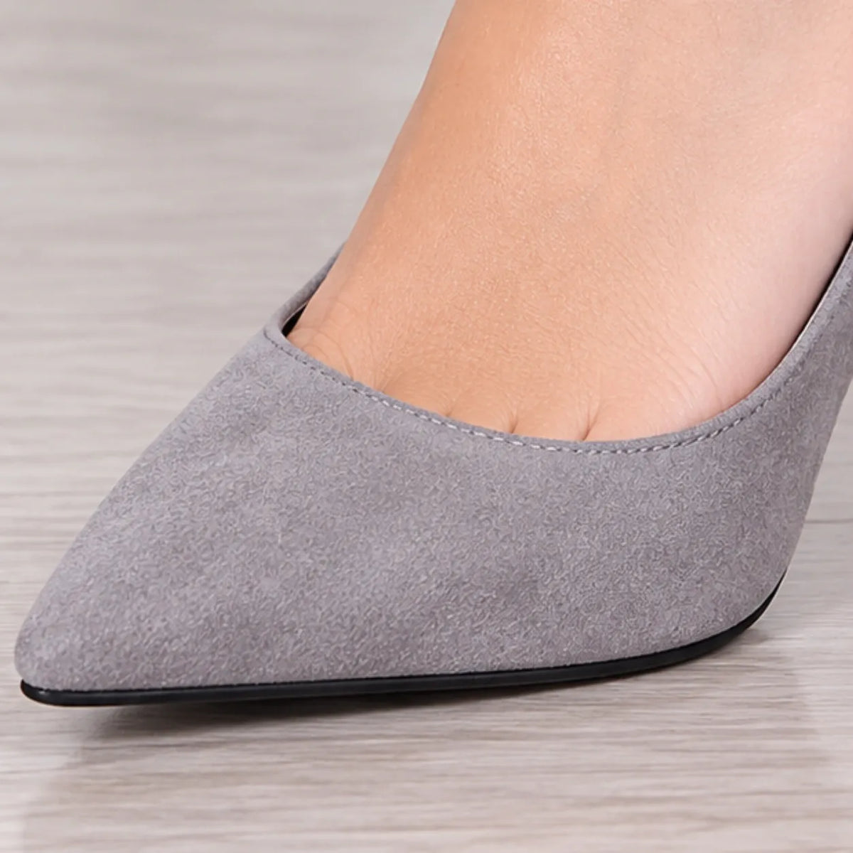 A close-up of a person's foot wearing a light gray suede pointed-toe pump on a light wood floor.