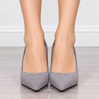 A pair of women's pointed-toe high heels in a light gray suede-like material, shown worn on a model's feet against a neutral indoor background with a light wood floor and white wall.